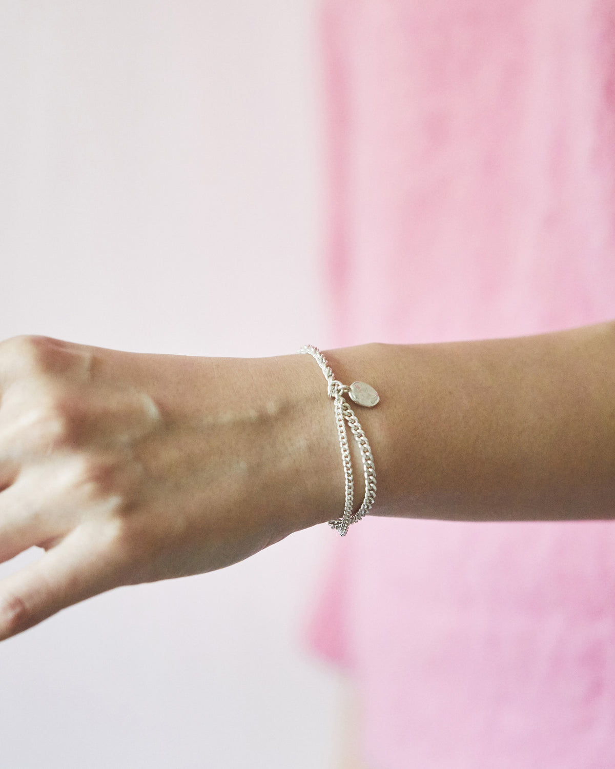 A close-up of a person's wrist wearing a delicate silver chain bracelet with a small oval charm, against a soft pink and white blurred background.