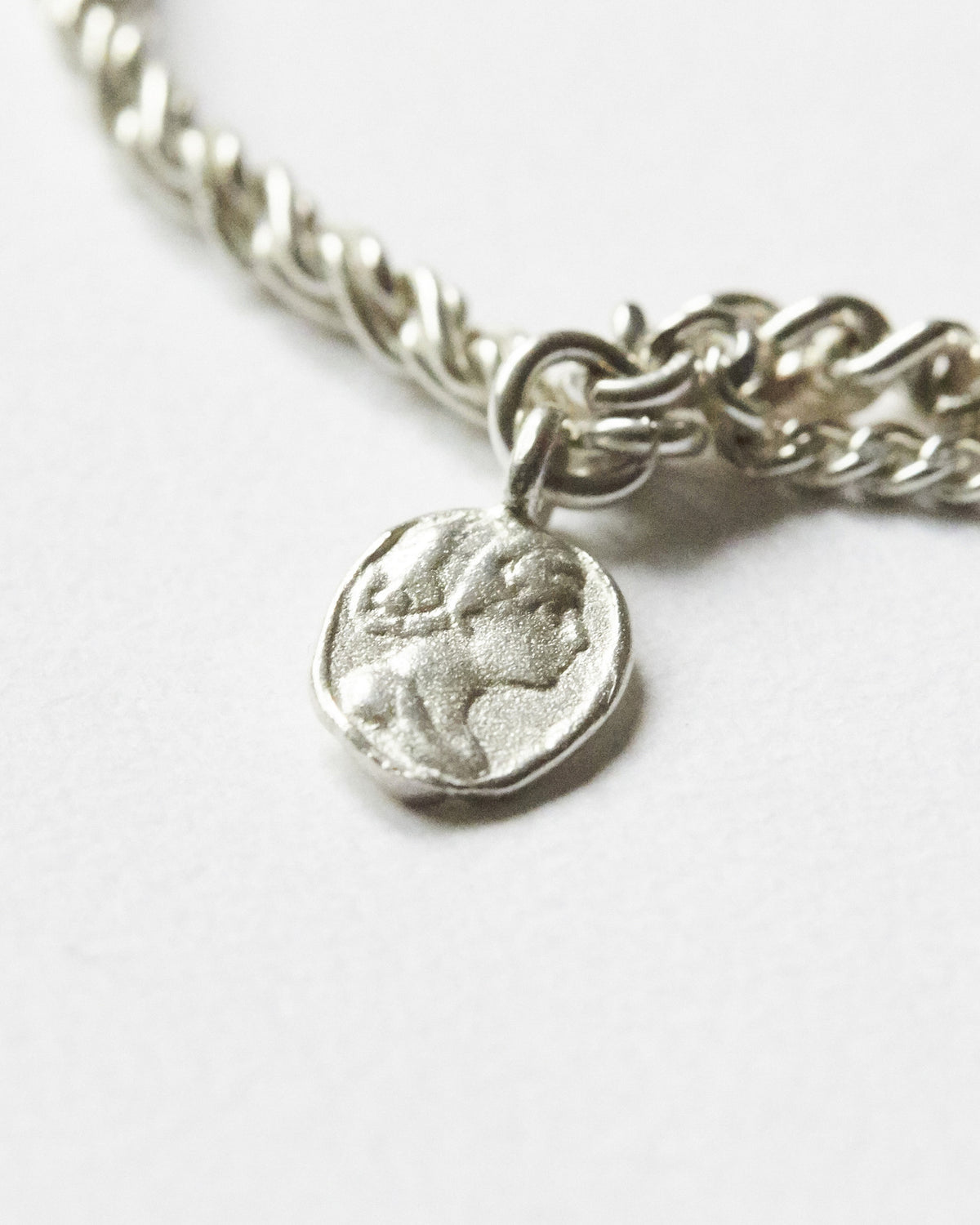 Close-up of a textured silver coin charm attached to a detailed chain bracelet, highlighting its irregular surface and handcrafted-inspired finish.