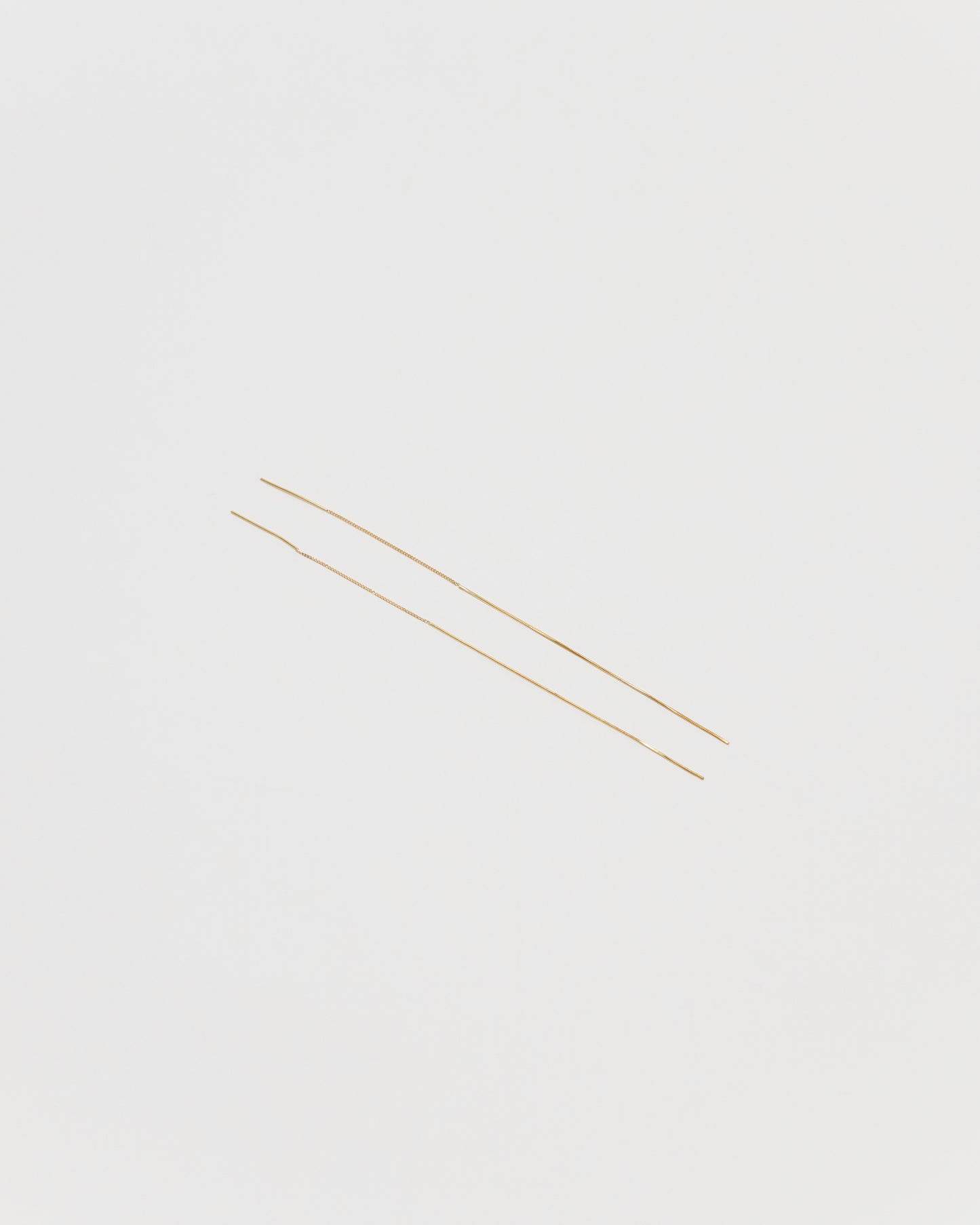Two extremely thin gold chain strands laid diagonally on a solid light gray background.