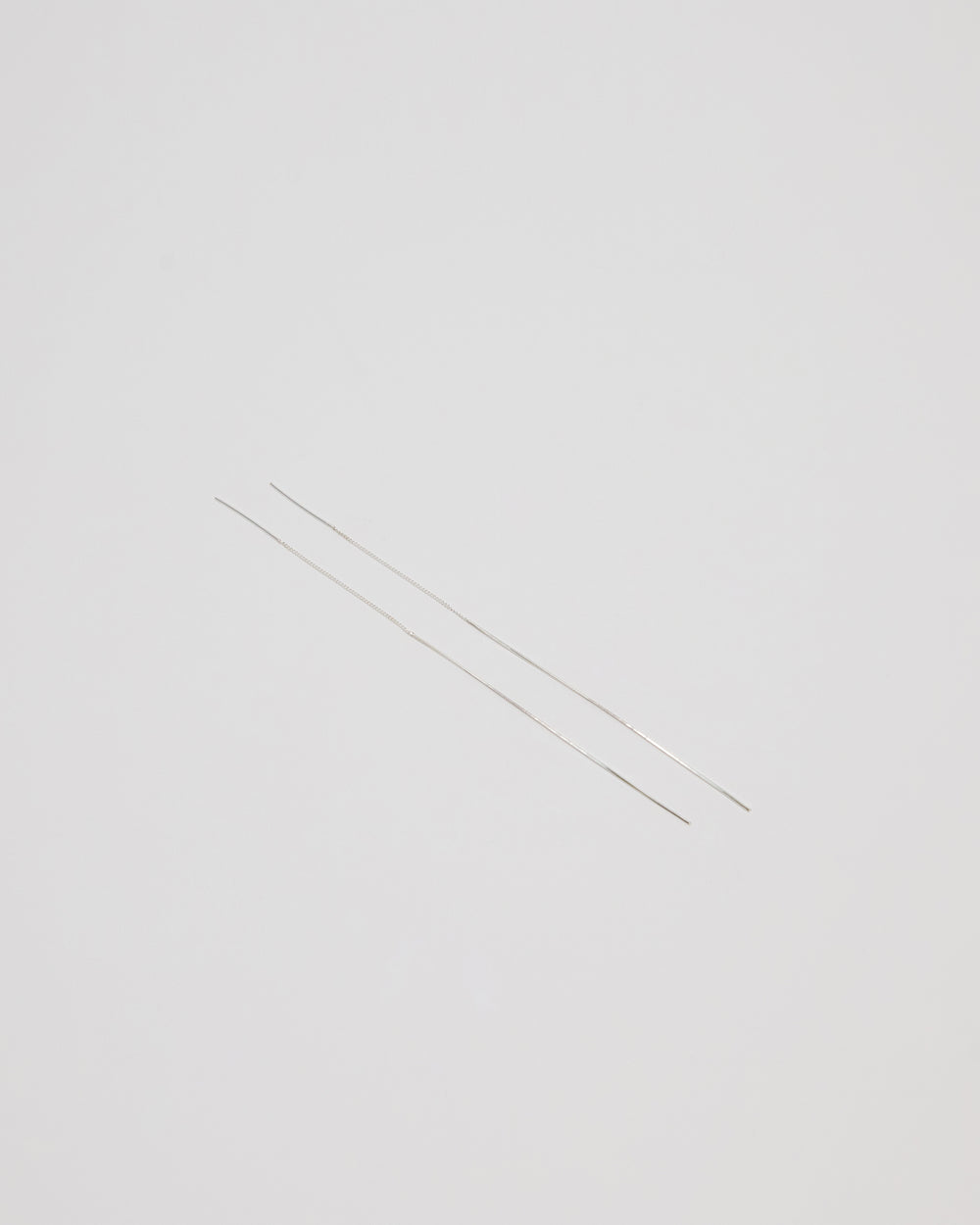 Two extremely thin silver metal rods, likely needle-like jewelry components or wire, laid diagonally on a solid light gray background.