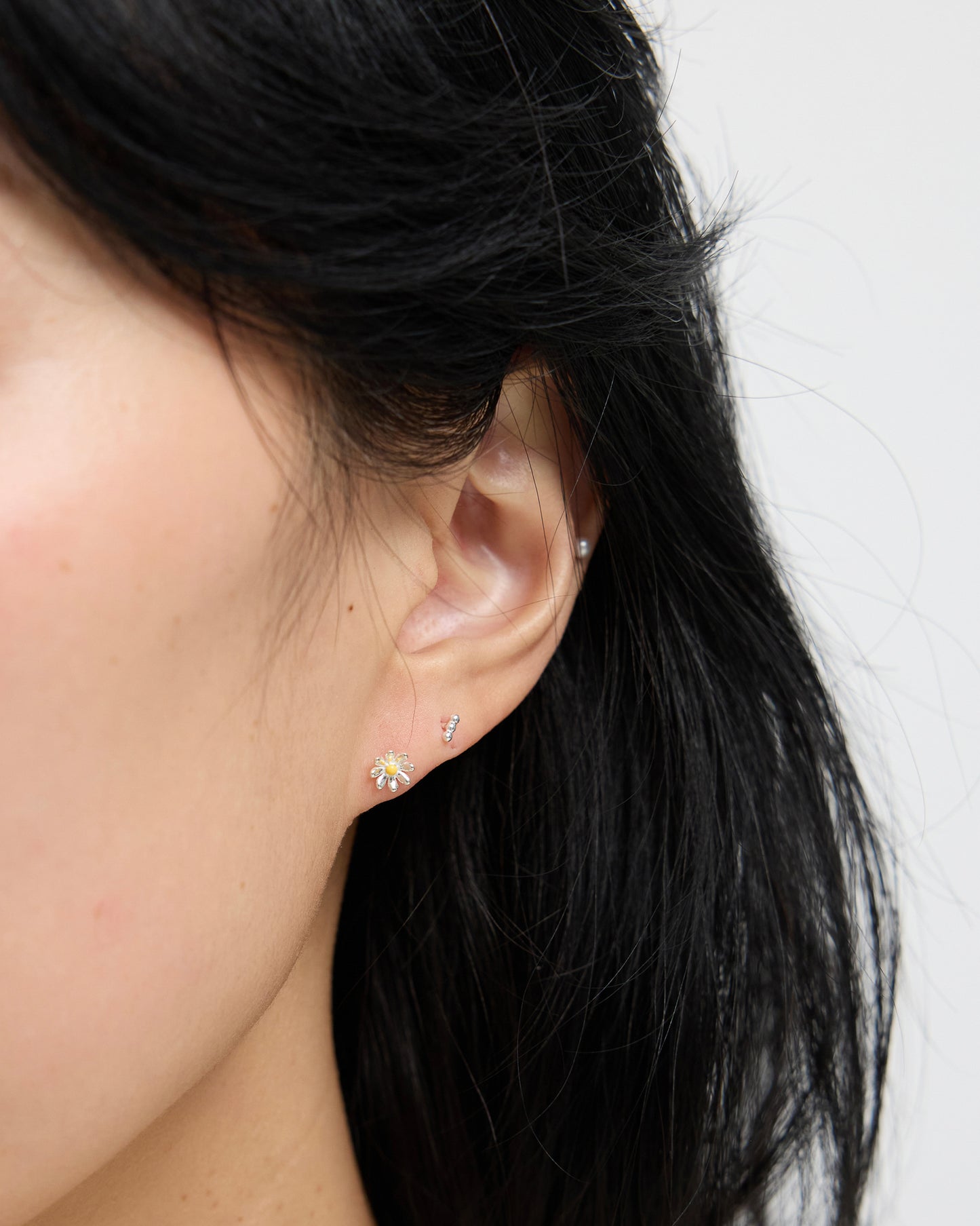 Close-up of an ear wearing a small silver daisy stud earring with a yellow enamel center, styled with an additional minimal bar stud above.