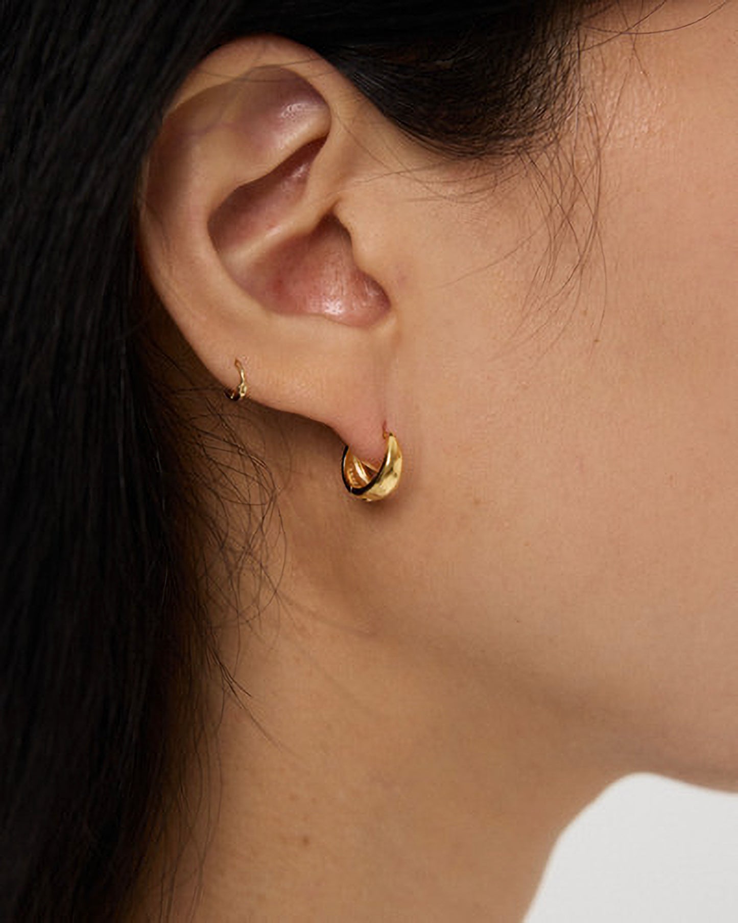 Close-up of an ear wearing a gold hoop earring with dark hair and white clothing.