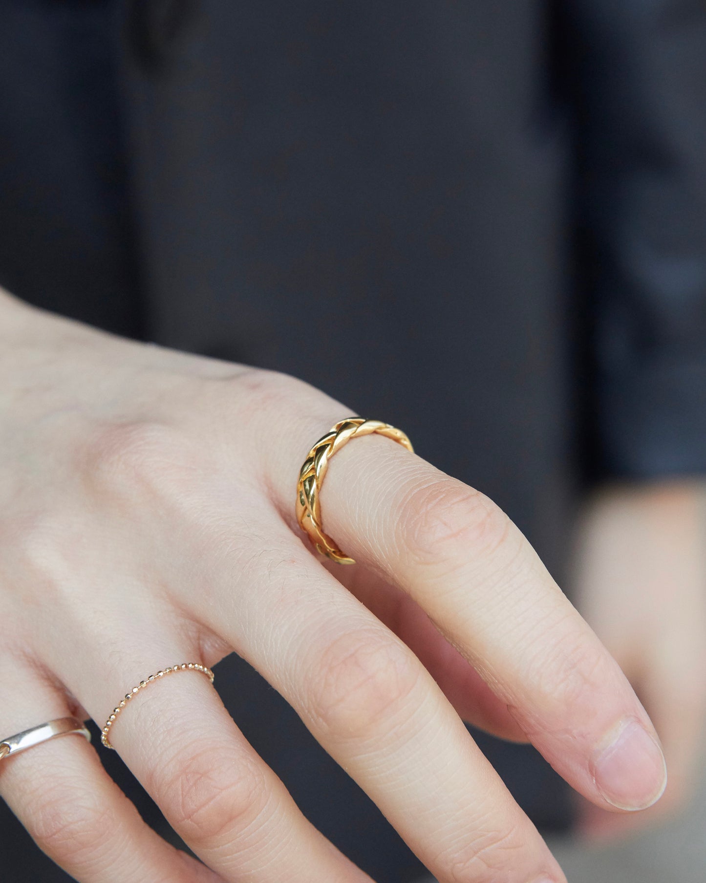 Gold braided ring styled solo on finger, creating a clean and warm contrast against a dark outfit.