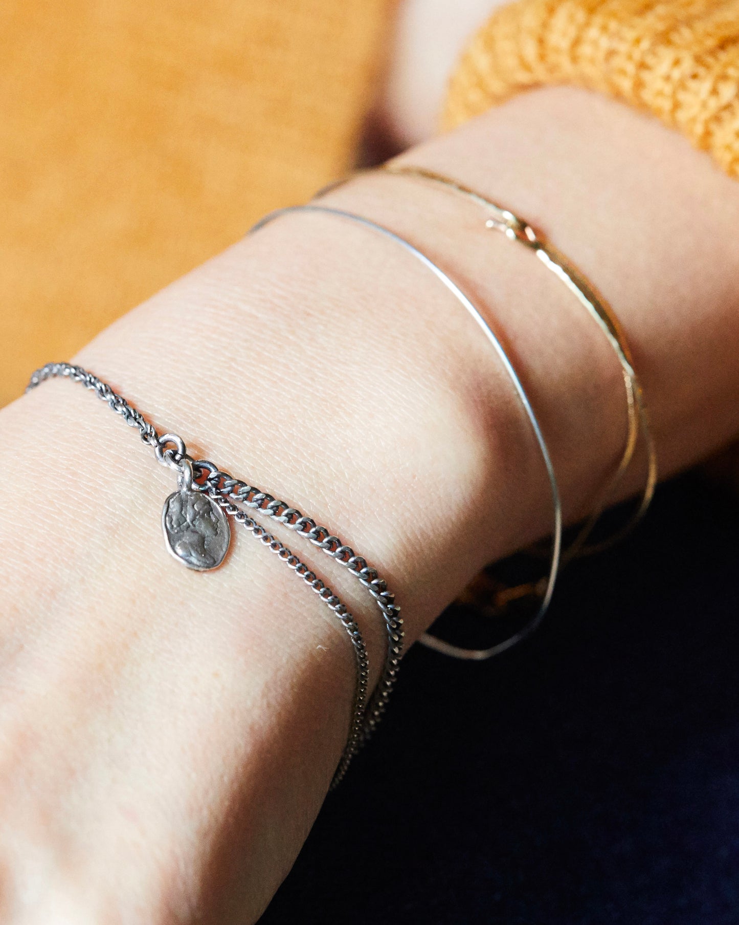 Silver charm bracelet worn on wrist, styled with mixed-metal bangles and mustard sweater