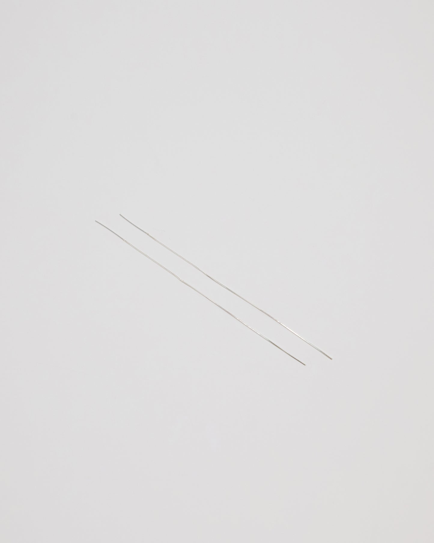Two extremely thin silver metal rods, likely needle-like jewelry components or wire, laid diagonally on a solid light gray background.