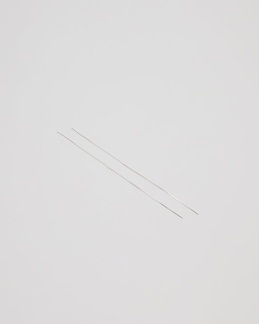 Two extremely thin silver metal rods, likely needle-like jewelry components or wire, laid diagonally on a solid light gray background.
