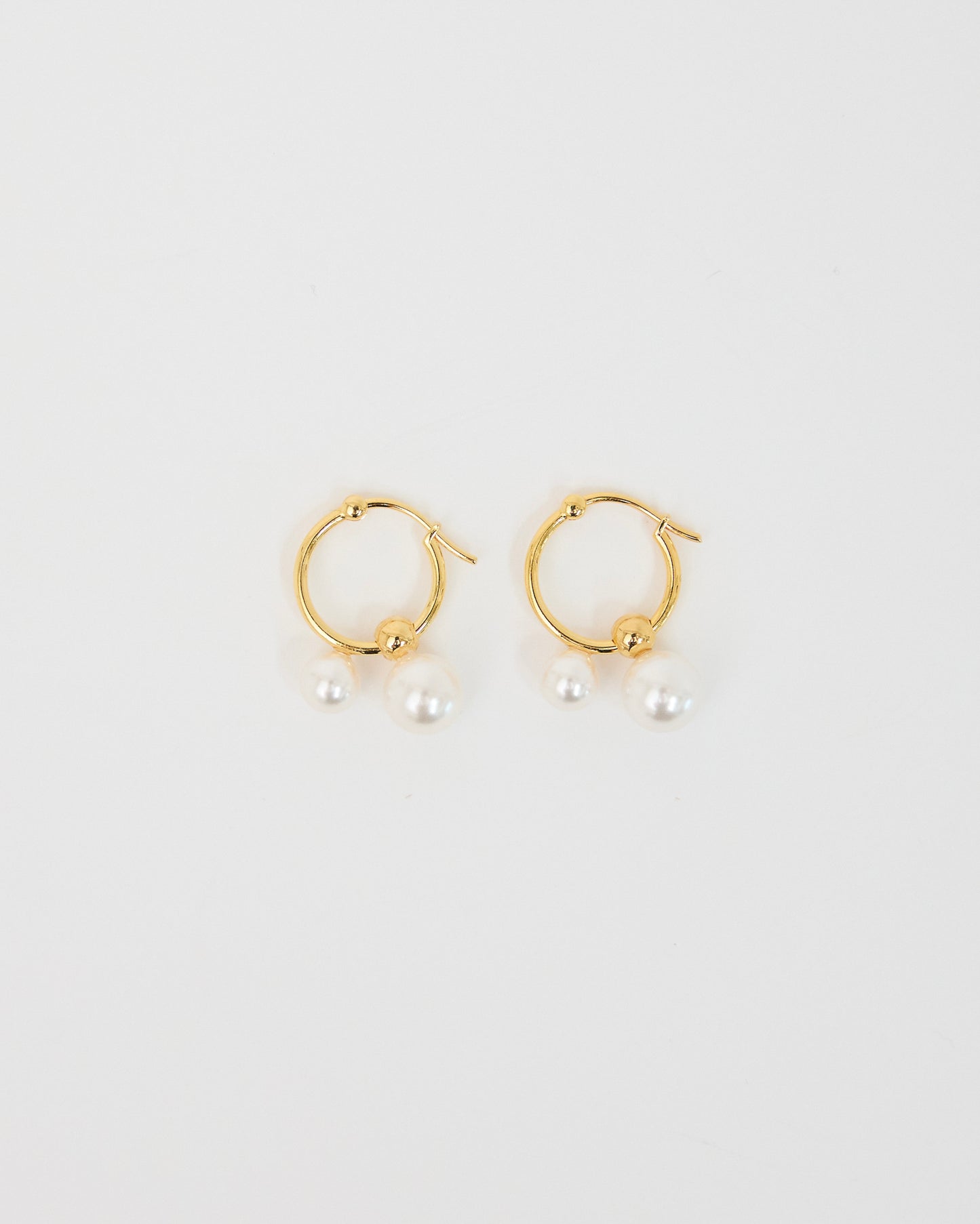 Pair of gold-tone hoop earrings with two white pearl drops placed on a soft white surface, photographed as a clean minimal product shot.