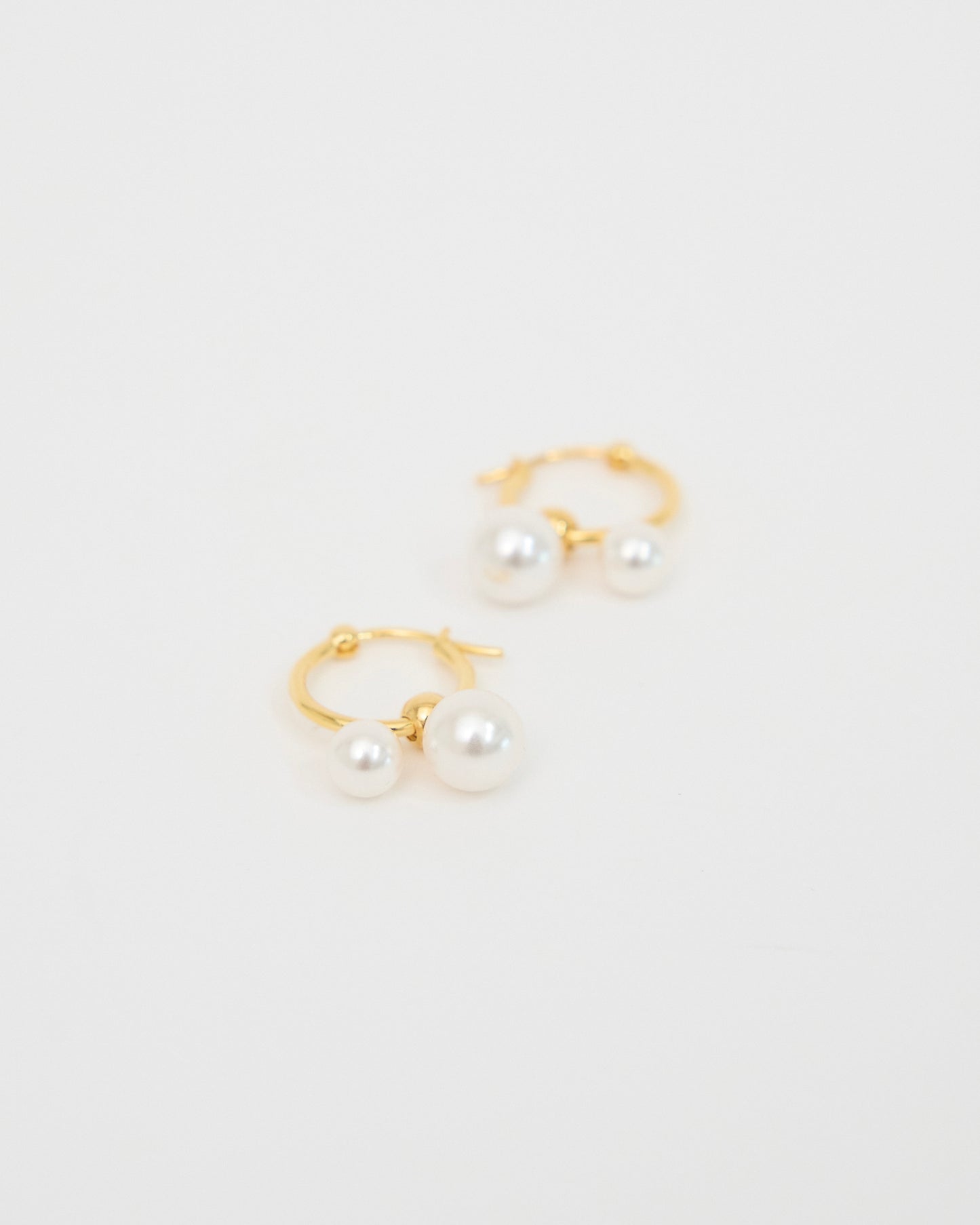 Pair of gold-tone hoop earrings with two white pearl drops placed on a soft white surface, photographed as a clean minimal product shot.